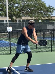 A male pickleball player wearing a black tank top, blue shorts, black leggings and visor demonstrates the two handed backhand shot on a pickleball court with a lush background and pickleball facility in the background.